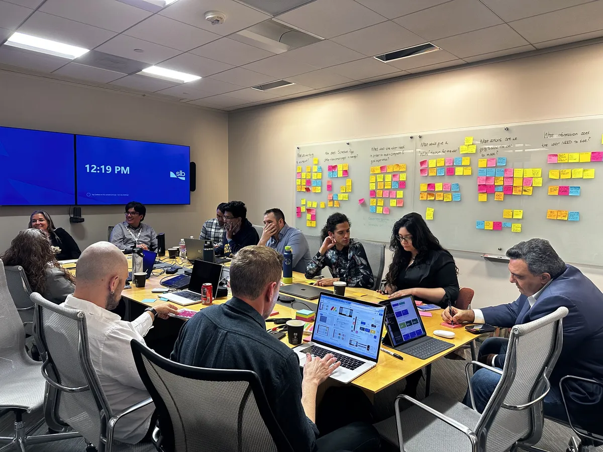 The whole team around a long table with laptops, the wall behind them covered in sticky notes under the workshop questions.