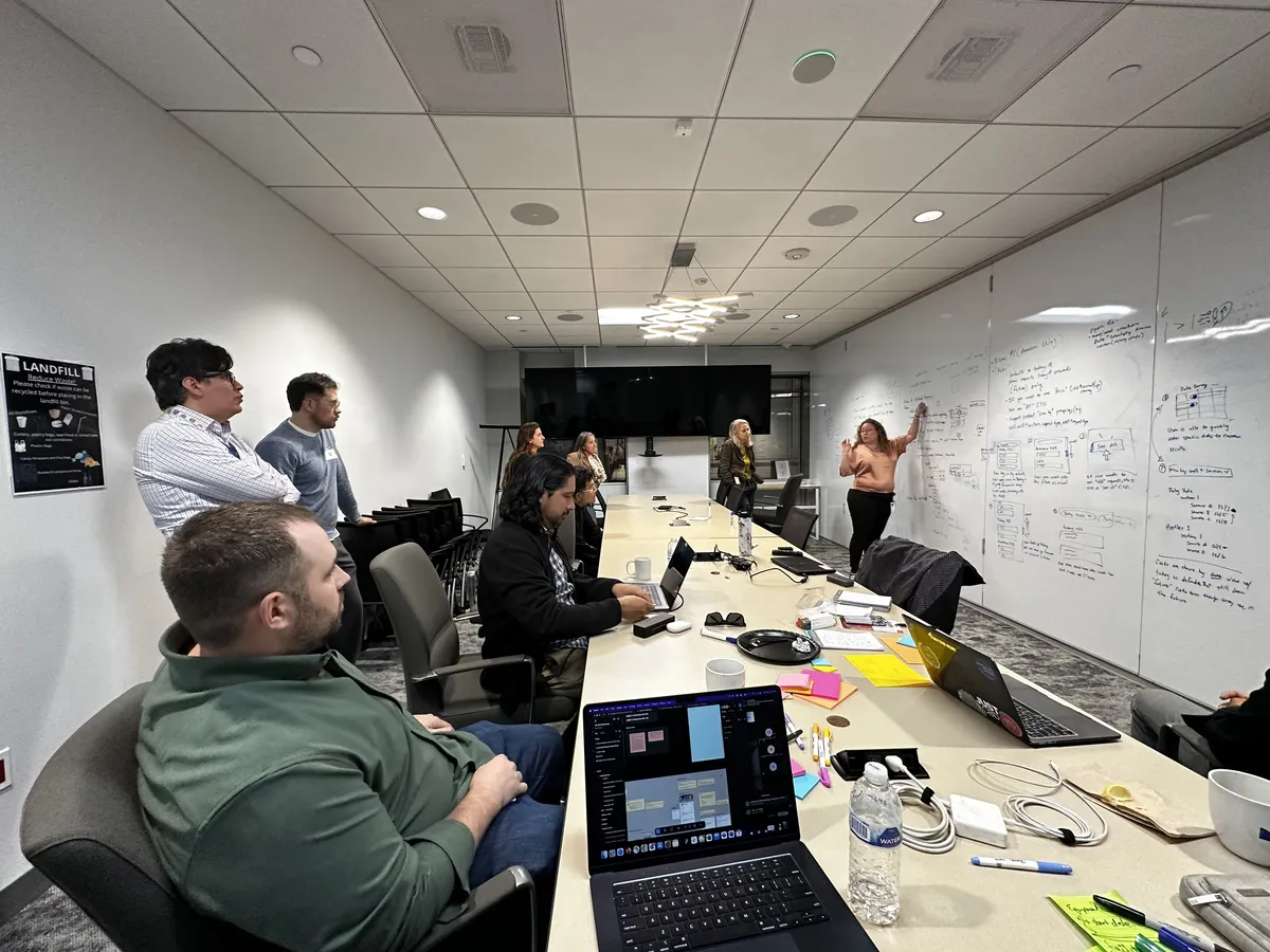 A long conference table, the team looking on while a designer sketches flows and screens on the whiteboard wall.
